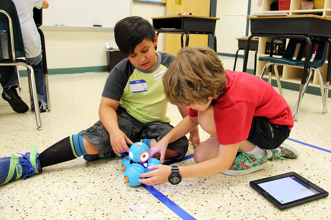 Two elementary students on a classroom floor programming a small blue robot beside a tablet